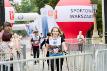 Kobieta z jasnymi włosami, ubrana na sportowo, idzie z kijkami do nordic walking. W tle stoi duży czerwono-biały balon z napisem „SPORTOWA WARSZAWA”. Po lewej stronie grupa osób bije brawo. Dalej widać uczestników biegu oraz bramę mety z logotypami sponsorów.