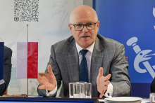 Mężczyzna w średnim wieku, w okularach - rektor WUM siedzi przy stole i gestykuluje rękami. Ma na sobie szary garnitur i niebieski krawat. 