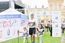 Mężczyzna trzyma za ręce dwoje dzieci. Wszyscy noszą koszulki z kolorowym nadrukiem i napisem „Bieg po nowe życie”. Stoją na zielonej trawie przed żółtym pałacem. W tle widać biały namiot z logo PZU oraz bramę startową z logotypami sponsorów. 