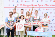 Osiem osób stoi na tle ścianki z napisem „28. Bieg po Nowe Życie – Warszawa 2023”. Dwie kobiety i dwóch mężczyzn noszą koszulki z napisem „Razem dla Transplantologii” i medalami na szyjach. Każda osoba trzyma medal w ręce. Na ściance widnieją logotypy sponsorów.