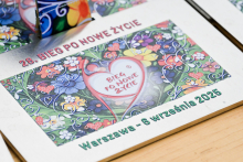 Plakat promuje 28. Bieg po Nowe Życie. W centrum widnieje czerwone serce z białym napisem „Bieg po Nowe Życie”. Tło tworzą kolorowe kwiaty i liście. Na górze znajduje się napis „28. BIEG PO NOWE ŻYCIE”, a na dole „Warszawa – 6 września 2025”.