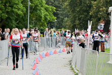Grupa kobiet i mężczyzn idzie trasą nordic walking na świeżym powietrzu. Uczestnicy noszą kolorowe koszulki z numerami startowymi. Niektórzy trzymają kijki. Trasa jest oznaczona pachołkami i otoczona barierkami. W tle widać drzewa, widzów i innych uczestników.
