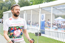 Mężczyzna ubrany w kolorową koszulkę z napisami „Bieg po nowe życie” i „Polska Transplantologia”. Trzyma kijki do nordic walking. Ma okulary przeciwsłoneczne na głowie i zegarek na lewym nadgarstku. W tle stoi biały namiot z przezroczystymi ścianami. W środku przebywają inni uczestnicy.