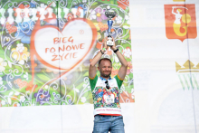 Na scenie mężczyzna w sportowym stroju. W rękach uniesionych nad głową trzyma puchar. Mężczyzna uśmiecha się. Za nim widać kolorowy baner. 