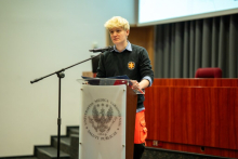 Student stoi przy przezroczystej mównicy z logo Uniwersytetu Medycznego w Warszawie. Ma na sobie ciemny sweter z emblematem oraz pomarańczowe spodnie