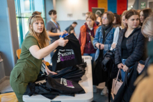 Grupa studentów stoi przy stole, na którym ułożone są czarne torby z napisem „Samorząd Studentów Warszawski Uniwersytet Medyczny”. Osoba w zielonym stroju podaje jedną z toreb osobie stojącej po prawej stronie.