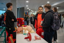 Grupa osób stoi przy stole, na którym znajdują się modele anatomiczne i sprzęt medyczny. Na pierwszym planie dwie osoby w strojach ratowników medycznych.
