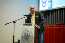 Mężczyzna w okularach, ubrany w ciemną marynarkę i białą koszulę, stoi przy mównicy z mikrofonem. 