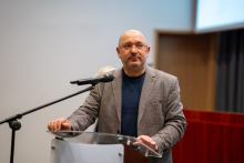 Mężczyzna w okularach, ubrany w ciemnoniebieską koszulkę i marynarkę w kratkę, stoi przy mównicy.