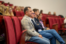 Na pierwszym planie mężczyzna w jasnej marynarce w kratę i niebieskich dżinsach siedzi z rękami opartymi na kolanach. Znajduje się w auli. Osoby obok niego również siedzą i patrzą przed siebie