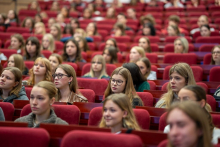 Duża grupa studentów siedzi w audytorium na czerwonych, wyściełanych krzesłach ustawionych w rzędach. 