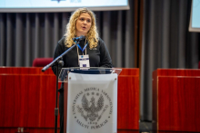 Młoda kobieta stoi za przezroczystą mównicą z logo Uniwersytetu Medycznego w Warszawie. Ma na sobie ciemną bluzę z napisem „WUM”