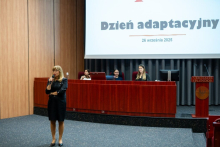 Kobieta stoi po lewej stronie sali konferencyjnej i przemawia do publiczności, trzymając mikrofon. Na ścianie za nimi widoczny jest duży ekran z napisem „Dzień adaptacyjny” 