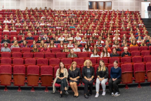 Duża grupa studentek i studentów siedzi w audytorium na czerwonych fotelach ustawionych w rzędach. W pierwszym rzędzie siedzi pięć osób