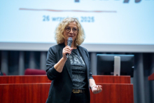 Kobieta o jasnych, kręconych włosach i w okularach stoi na tle sali konferencyjnej, trzymając mikrofon w prawej ręce