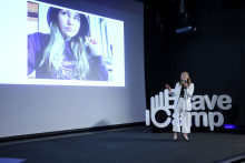 Magdala Halska. She stands in front of a bright screen with the ConsentHub logo. She holds a microphone in her right hand. She has long, light hair. She wears a black shirt and black pants. Behind her, you can see a padlock icon and a checklist.