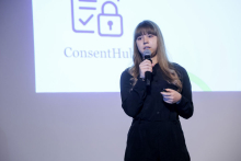 Kaja Tyszkiewicz. A young woman with long light brown hair stands on a brightly lit stage and speaks into a microphone. She is wearing a black shirt and pants. She looks straight ahead. Behind her is a white screen with the ConsentHub logo featuring a padlock icon and a checklist. 