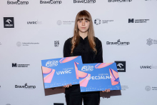 Kaja Tyszkiewicz. A young woman with long, straight hair stands in front of a wall with the logos of BraveCamp, Warsaw University of Technology, and SWPS University. She is wearing black clothes. She is holding two blue posters with the words “UWRC award” and “SPECIAL award.”