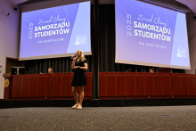 Kobieta stoi na scenie w sali konferencyjnej i trzyma mikrofon. W tle widoczne są dwie duże projekcje z napisem „Zarząd Główny Samorządu Studentów 2025 Dni Adaptacyjne”