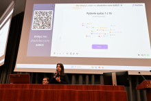 Młoda kobieta stoi na scenie za stołem prezydialnym. W tle na dużym ekranie wyświetlona jest strona internetowa z quizem. 