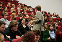 Mężczyzna w jasnej marynarce stoi w sali wykładowej i trzyma mikrofon, rozmawiając z jedną z kobiet siedzących na widowni. 