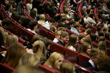 Duża grupa młodych kobiet i mężczyzn siedzi w audytorium na czerwonych, wyściełanych krzesłach ustawionych w rzędach. Wszyscy są zwróceni w stronę sceny, której nie widać na zdjęciu