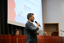 Mężczyzna w marynarce przemawia do mikrofonu, stojąc na tle dużego ekranu projekcyjnego 