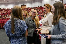 Grupa pięciu kobiet stoi w audytorium pełnym ludzi. W tle widoczne są rzędy czerwonych krzeseł, na których siedzą uczestnicy wydarzenia. Kobiety na pierwszym planie są ubrane formalnie i rozmawiają ze sobą.