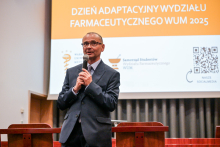 Mężczyzna w garniturze przemawia do mikrofonu w sali wykładowej. W tle widoczny jest ekran z napisem „Dzień adaptacyjny Wydziału Farmaceutycznego WUM 2025”. 