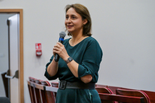 Kobieta w zielonej sukience z długimi rękawami i czarnym paskiem w talii stoi w pomieszczeniu, trzymając mikrofon w prawej ręce. 