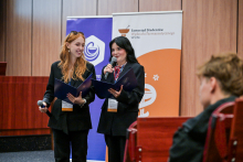 Dwie młode kobiety stoją na scenie w auli. Ubrane są na ciemno. W tle widoczne są dwa roll-upy: jeden z logo i napisem „Samorząd Studentów Wydziału Farmaceutycznego WUM”,