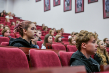 Grupa młodych kobiet i mężczyzn siedzi w audytorium na czerwonych, tapicerowanych krzesłach ustawionych w rzędach.