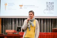 Kobieta trzyma mikrofon i stoi na tle ekranu projekcyjnego w auli.