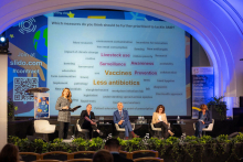 Five people on stage. They are participants in a thematic session. Four people are sitting, one is standing and holding a microphone. Behind them is a multimedia screen.