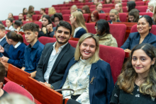 Cztery osoby siedzą w czerwonych fotelach w sali wykładowej. Dwie kobiety i dwóch mężczyzn uśmiechają się do kamery. W tle widać kilkanaście osób w eleganckich ubraniach, takich jak marynarki i koszule. Rozmawiają ze sobą i również siedzą.
