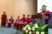 Siedem osób uczestniczy w uroczystości akademickiej. Sześć osób w czerwono-czarnych togach i biretach siedzi na scenie. Jedna osoba stoi przy mównicy z mikrofonem i przemawia. W tle widać drewniany panel i ekran z tekstem. Na pierwszym planie leżą białe kwiaty z zielonymi liśćmi.