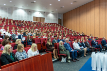 W sali wykładowej z czerwonymi fotelami siedzi kilkadziesiąt osób. W pierwszych rzędach znajdują się kobiety i mężczyźni w formalnych strojach. Za nimi siedzą młodzi ludzie, prawdopodobnie studenci, ubrani elegancko. 