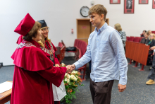 Młody mężczyzna w jasnoniebieskiej koszuli ściska dłoń kobiety w czerwonej todze i birecie. Obok stoi druga kobieta w czarnej todze i birecie, trzyma torbę. W tle widać czerwone krzesła, zegar, portrety na ścianie i kilka siedzących osób. 