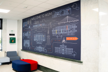 Jasne wnętrze z marmurowymi ścianami i sufitem podwieszanym. Na ścianie widnieje duża grafika z mapą kampusu WUM i napisem „WELCOME TO WUM”. Mapa pokazuje budynki, ścieżki i pomarańczowy prostokąt z napisem „WELCOME CENTER”. Przed grafiką stoją dwa siedziska: niebieskie i czerwone. 