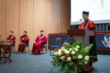 Cztery osoby w togach na scenie auli. Trzy siedzą. Jedna osoba - dziekan wydziału stoi przy mównicy i przemawia. Na mównicy widnieje herb uczelni. Na pierwszym planie stoi duży bukiet z białymi różami i czerwonymi owocami. W tle widać fragment ekranu z napisem Facultas Pharmaceutica.