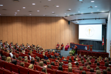 Aula w czasie uroczystości. Na scenie cztery osoby ubrane w togi akademickie, trzy siedząm jedna stoi przy mównicy. Na czerwonych fotelach siedzi kilkadziesiąt osób, wszystkie patrzą w stronę sceny.