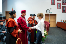 Studentka, dziekan, prodziekanki i prorektor stoją na środku auli. Studentka i prorektor podają sobie ręce. Wszyscy się uśmiechają.