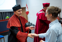 Prodziekanka ściska dłoń studentki, ubranej w niebieską koszulę. Prodziekanka w lewej dłoni trzyma srebrną przypinkę. W tle widać inne osoby.