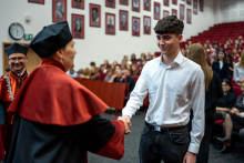 Prodziekanka ściska dłoń studenta, ubranego formalnie. W tle widać inne osoby.
