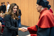 Prodziekanka ściska dłoń studentki, ubranej elegancko w czarną marynarkę. W tle widać inne osoby.