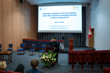  Na scenie, za mównicą stoi dr Białek. Ma na sobie czarną togę akademicką. Przemawia.  Na ekranie za nim widnieje slajd z tytułem prezentacji: ‘Dlaczego uczulenie na ten sam alergen może mieć odmienny przebieg kliniczny u różnych pacjentów?’.