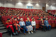 Audytorium wypełnione osobami w togach i biretach podczas ceremonii ukończenia studiów; w pierwszym rzędzie osoby w strojach akademickich i formalnych