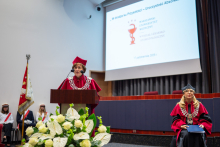 Osoba w stroju akademickim przemawia przy mównicy podczas uroczystości dyplomowania absolwentów.