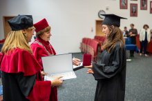 Absolwentka w todze odbiera dyplom i medal od dwóch osób w czerwonych togach.