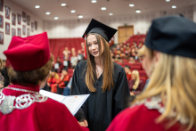Osoba w todze odbiera dyplom od dwóch osób w czerwonych togach podczas ceremonii wręczenia dyplomów.
